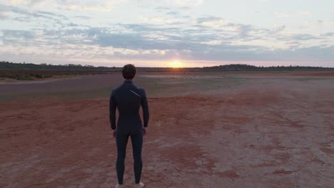 Male-model-wearing-wetsuit,-looking-at-camera-then-turning-round-to-look-towards-sunrise