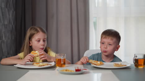 siblings sitting together at dining table eating pizza with berry juice in glass cups smiling at each other during cozy indoor meal with soft natural light filtering through