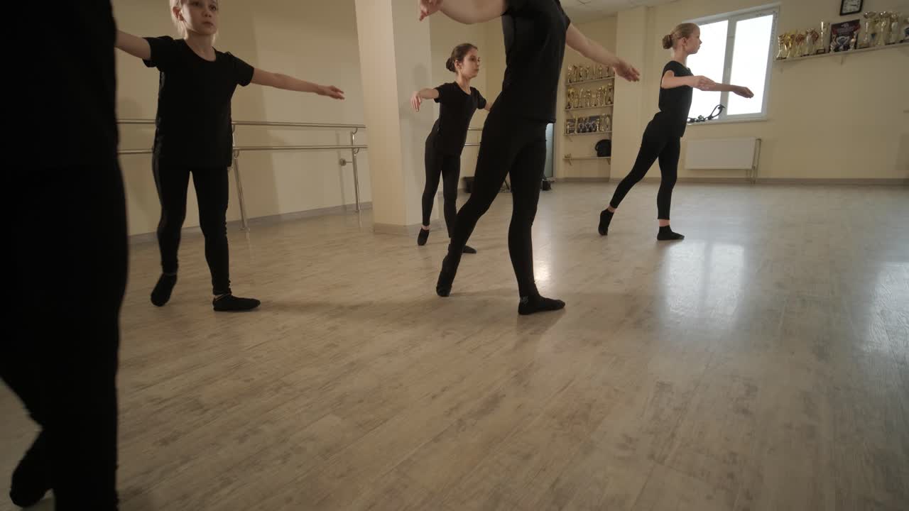 un grupo de jóvenes estudiantes de ballet con ropa de baile negra practicando posiciones en un amplio estudio de ballet con pisos de madera y barras montadas en la pared. expresiones enfocadas y movimientos sincronizados.