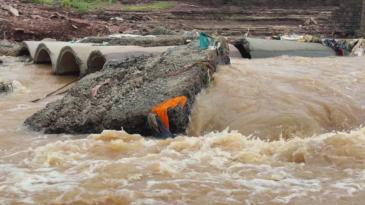 Static shot showing a heavly damaged culvert or a small bridge partially submerged by the powerful, muddy brown water during the flooding