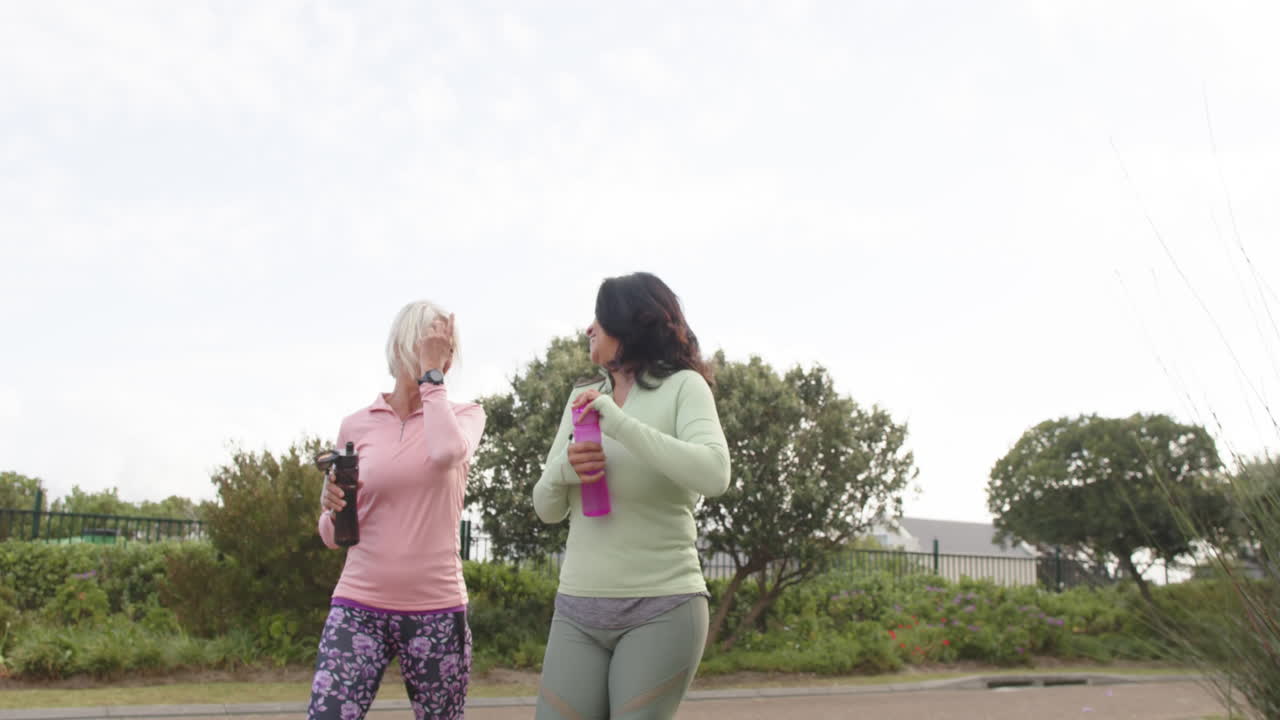 dos mujeres mayores diversas caminando con botellas de agua hablando en un día soleado, cámara lenta, espacio de copia