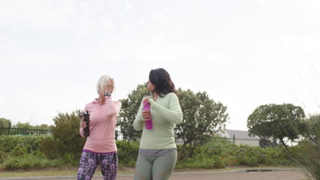 two diverse senior women walking with water bottles talking on sunny day, slow motion, copy space