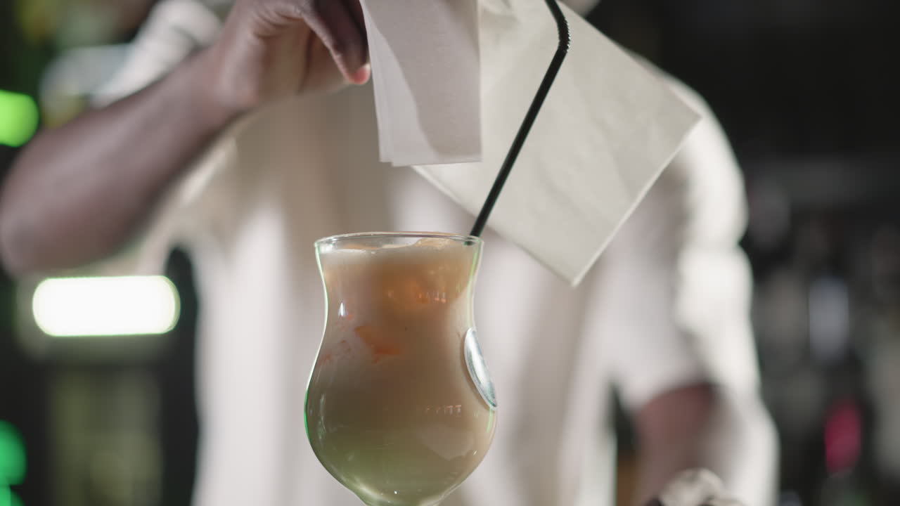 Bartender placing straw in freshly made cocktail, serving drink in elegant glass. Behind, illuminated Heineken neon sign and bar shelves, enhancing lively bar atmosphere