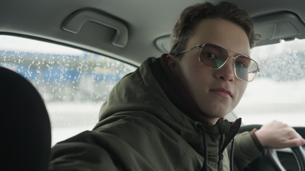 young driver in winter jacket and sunglasses sits behind steering wheel inside parked vehicle, turns around with subtle smile, looking directly at camera with blurred snowy background outside window