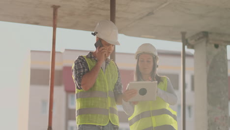 Supervisor-De-Un-Edificio-En-Construcción,-Un-Hombre-Discutiendo-Con-Una-Ingeniera-Diseñadora-Sobre-El-Progreso-De-La-Construcción-Y-Examina-Un-Plan-De-Construcción.