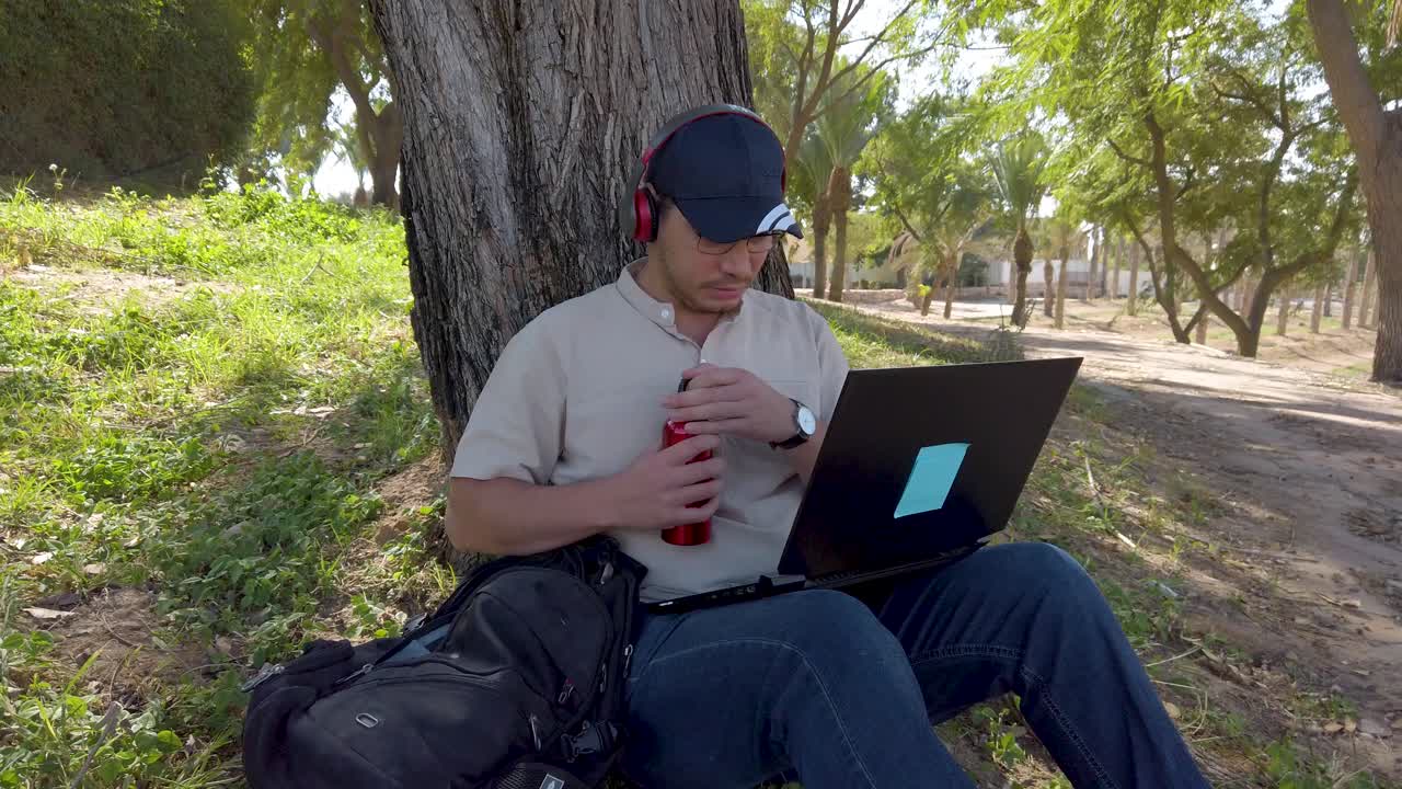 Man with cap using laptop in park. The scene is peaceful and relaxing, enjoying his time outdoors.