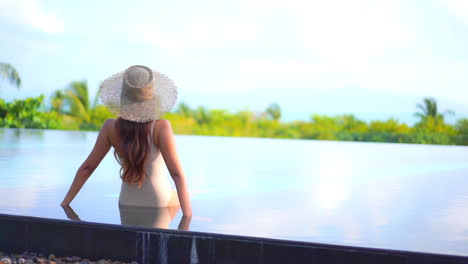 Sexy-Frau,-Die-Am-Rand-Des-Schwimmbades-In-Einem-Exotischen-Hotel-In-Hawaii-Sitzt-Und-Weiße-Monokini-Und-Sonnenhut-Trägt-Und-Auf-Tropische-Palmen-Blickt,-Rückansicht-In-Zeitlupe-Handheld