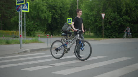 niño con pantalones grises y camiseta negra camina en su bicicleta a través de un cruce de peatones, con otros ciclistas visibles en el fondo, se ven varias señales y un coche en movimiento alrededor, con árboles
