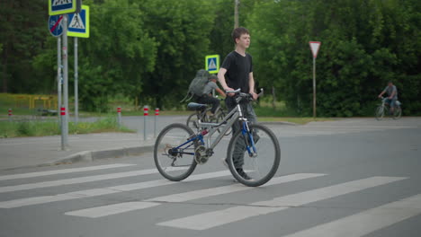 boy in grey trousers and black top walks his bicycle across a pedestrian crossing, with other cyclists visible in the background, several signposts and a moving car are seen around, with trees