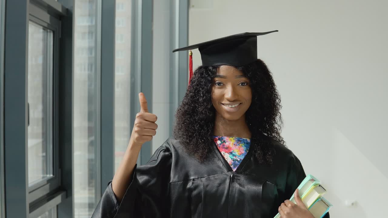una joven afroamericana graduada de la universidad se para con libros de texto en una mano y muestra el gesto bueno con el dedo gordo hacia arriba cerca de una gran ventana moderna con marcos negros.