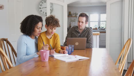 el padre trabaja en la computadora portátil mientras la madre ayuda al hijo con la tarea en la mesa de la cocina