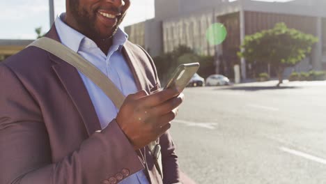 Sección-Media-De-Un-Hombre-Afroamericano-En-La-Ciudad,-Parado-Al-Sol-Usando-Un-Teléfono-Inteligente