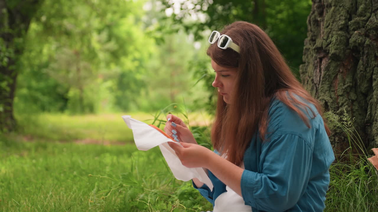 Una mujer con gafas sentada en un parque borda cuidadosamente una tela con un bastidor y una aguja, rodeada de árboles verdes y hierba desenfocados, con una suave luz del día que resalta el ambiente tranquilo de verano.