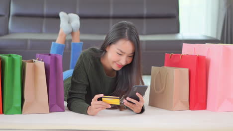 attractive asian girl lying on a floor surrounded with shopping bags using her credit card for shopping online, type in card information using a smartphone