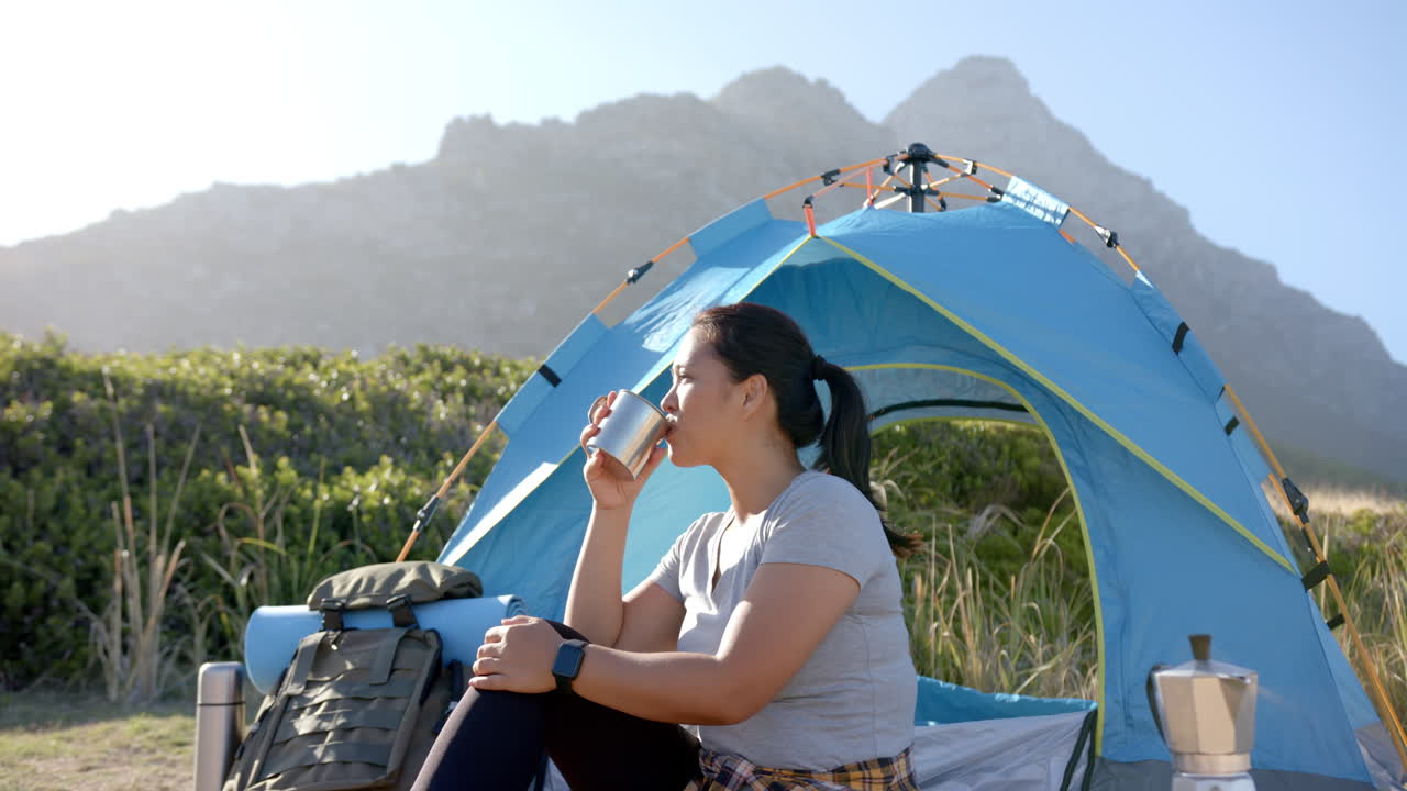 Camping in mountains, woman drinking from cup near blue tent with backpack