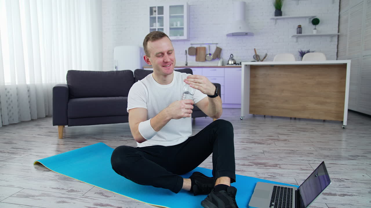 Handsome young sportsman doing exercises on quarantine. Lively athletic active man using his laptop and drinking water while training indoors