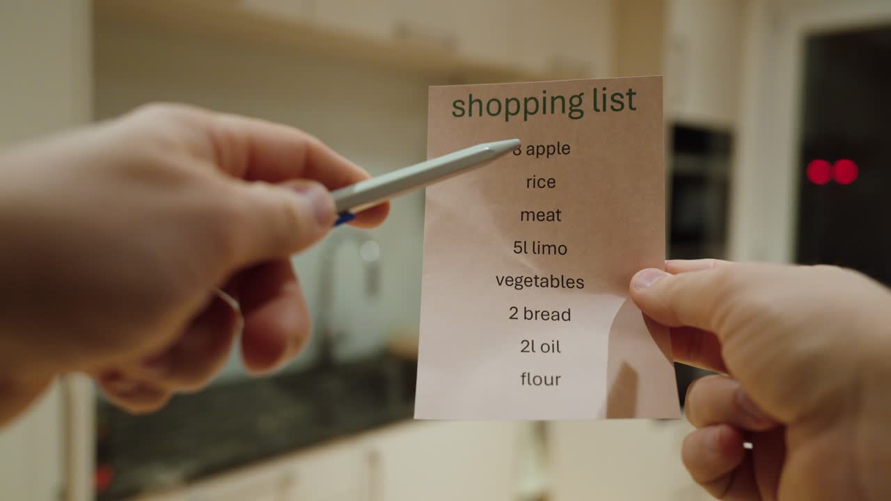 Närbild på händer som håller en inköpslista för mat i en köksmiljö. Koncept med måltidsplanering, hushållsorganisation, budgetering och daglig matlagningsrutin.