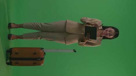 full body of asian businesswoman traveler with luggage taking note on a tablet while standing in the green screen background studio, during sunset time