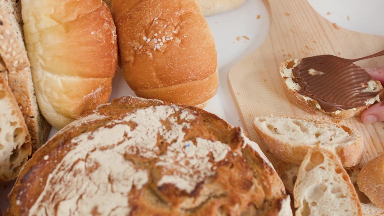 Close-up of hands spreading chocolate hazelnut cream on fresh baguette slices among assorted bakery breads.