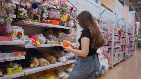 rear view of woman in black top holding and inspecting vibrant orange plush purse with green details in well-lit mall, she carefully examines item while browsing shelves filled with colorful toys