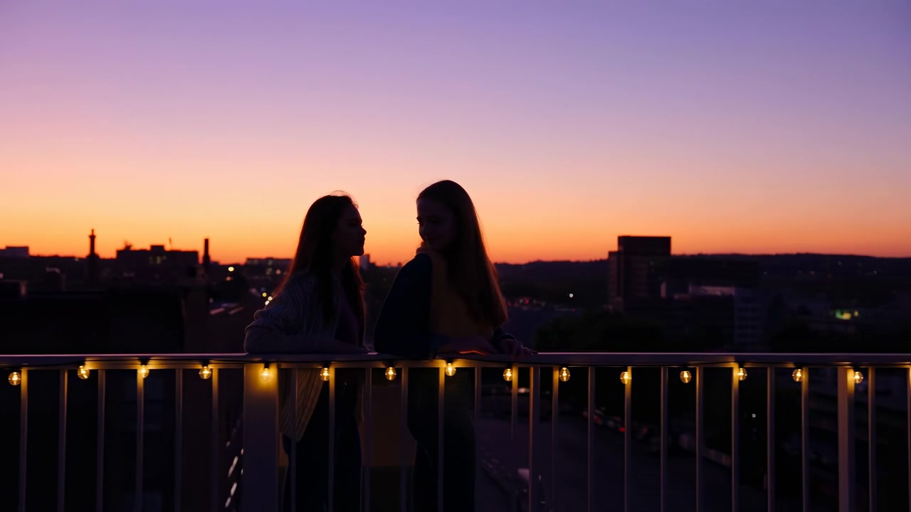 Two silhouetted women lean on rooftop railing facing each other at sunset, framed by city skyline, warm orange light, violet sky, soft string lights, quiet urban mood, with silhouette, women, rooftop