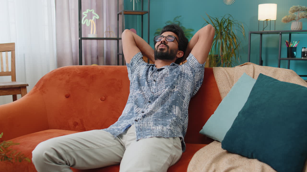 Tired indian man lying down on comfortable couch taking a rest daytime nap relaxing falling asleep
