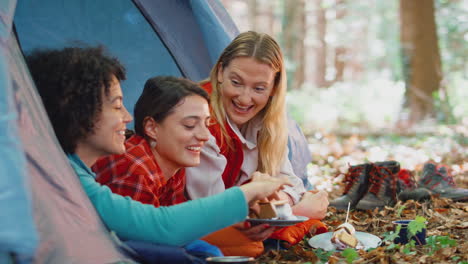 group of female friends on camping holiday in forest lying in tent eating s'mores