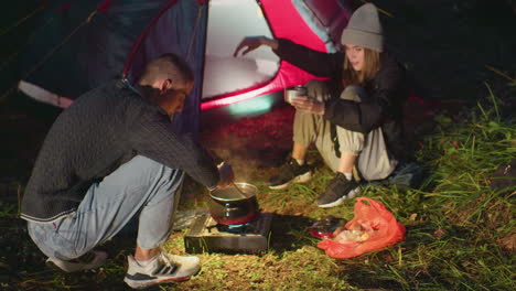 night camping scene with man stirring pot on portable gas stove while woman warms hands over hot bowl near tent entrance illuminated by lantern glow creating cozy retreat on damp grass