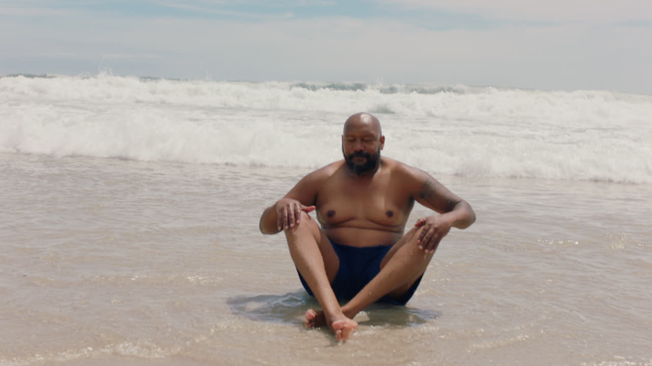 hombre afroamericano divertido sentado en la playa con las olas salpicando divirtiéndose en un cálido día de verano disfrutando de las vacaciones de verano 4k