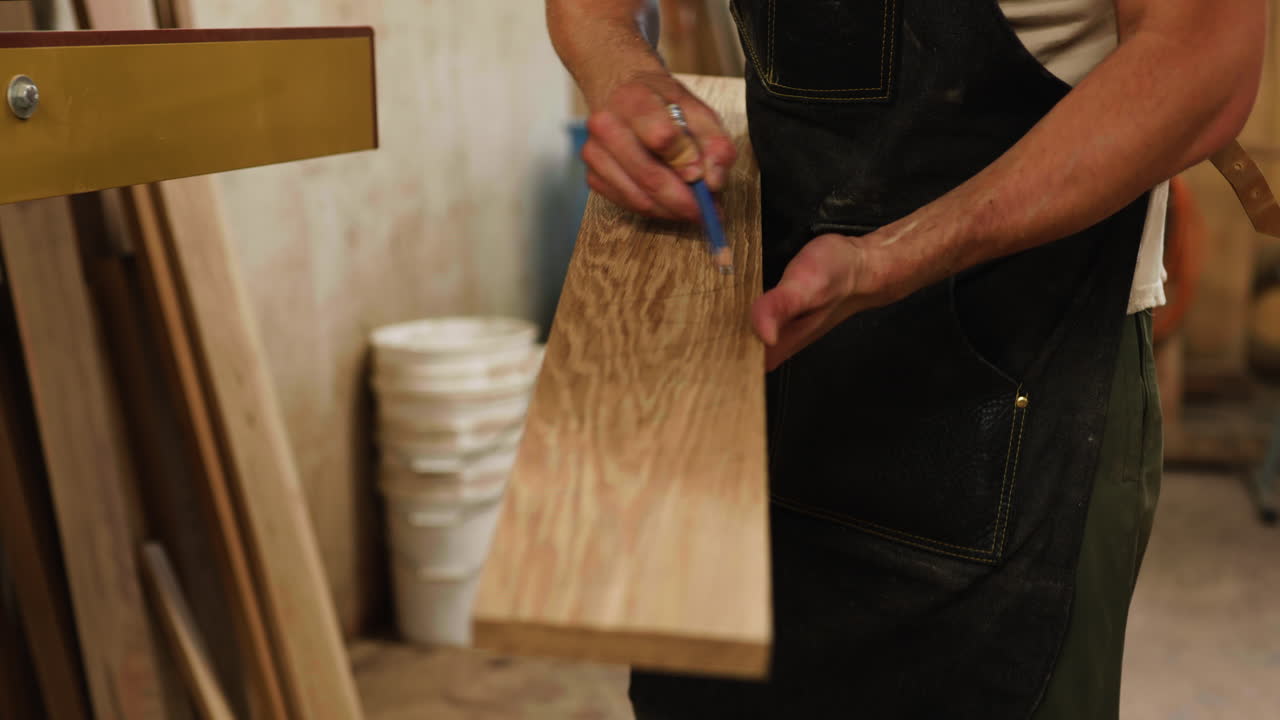 A skilled worker uses a pencil to carefully outline a design on a piece of oak wood in a well-organized workshop filled with tools and materials.