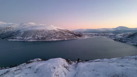 Slow-panning-aerial-shot-on-the-peak-of-a-snowy-mountain-surrounded-by-a-large-lake-and-snow-capped-mountains-during-winter-in-Ersfjordvegen-Norway