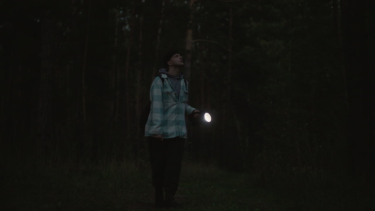 Excursionista caucásico en un bosque nocturno con una linterna que proyecta un haz de luz sobre un sendero cubierto de musgo, ambiente tenso de oscuridad y sombras, mirando hacia el cielo entre los troncos, ambiente de supervivencia aislado con un tenue resplandor y firme