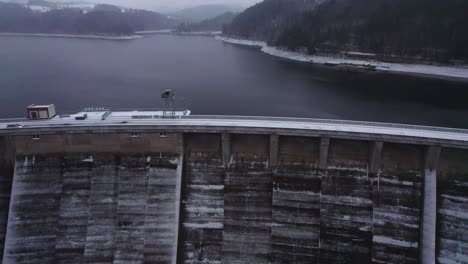 Vista-Aérea-Inclinada-Hacia-Arriba-Del-Embalse-Desde-Encima-De-La-Presa-De-Arco-De-Hormigón-Cubierta-Con-Una-Gruesa-Capa-De-Nieve