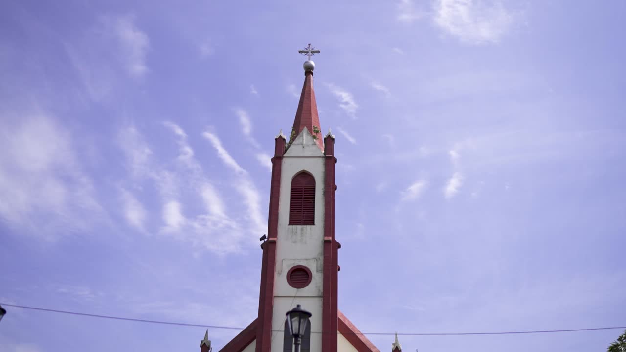 A low-angle shot of the historic Catholic church in Gobernador Roca, Misiones, Argentina. The red-roofed steeple stands tall against a beautiful blue sky with wispy clouds