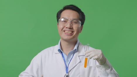 close up ofasian man scientist with orange liquid in the test tube smiling and showing thumbs up gesture to the camera while standing on the green screen background in the laboratory