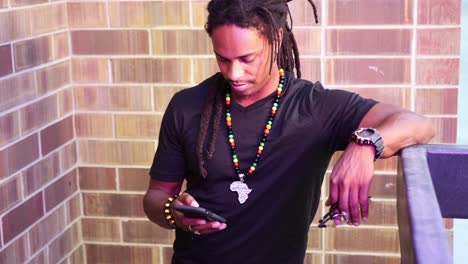 African-American-man-with-dreadlocks-leaning-against-railing-on-his-phone,-bobbing-his-head-to-music,-then-interacting-with-camera