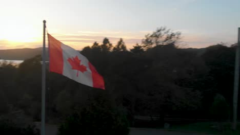 Toma-Aérea-Semi-orbital-De-Una-Bandera-De-Canadá-En-Un-Poste-Ondeando-Al-Viento-Con-Una-Impresionante-Puesta-De-Sol-Detrás-De-La-Línea-De-árboles