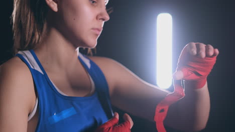 Young-beautiful-woman-wrapping-hands-with-black-boxing-wraps-in-dark-room.-Slowmotion-shot.-steadicam-shot