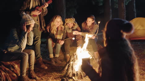 Amigos-Felices-Sentados-Junto-A-Una-Fogata-Joven-Tocando-Ukelele-Cantando-Disfrutando-De-La-Noche-Juntos-Acampando-En-El-Bosque-Por-La-Noche