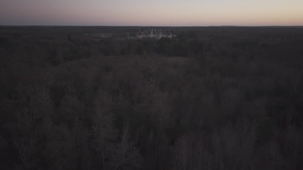 En vid flygbild visar den avlägsna silhuetten av Château de Chambord över mörka fält i skymningen
