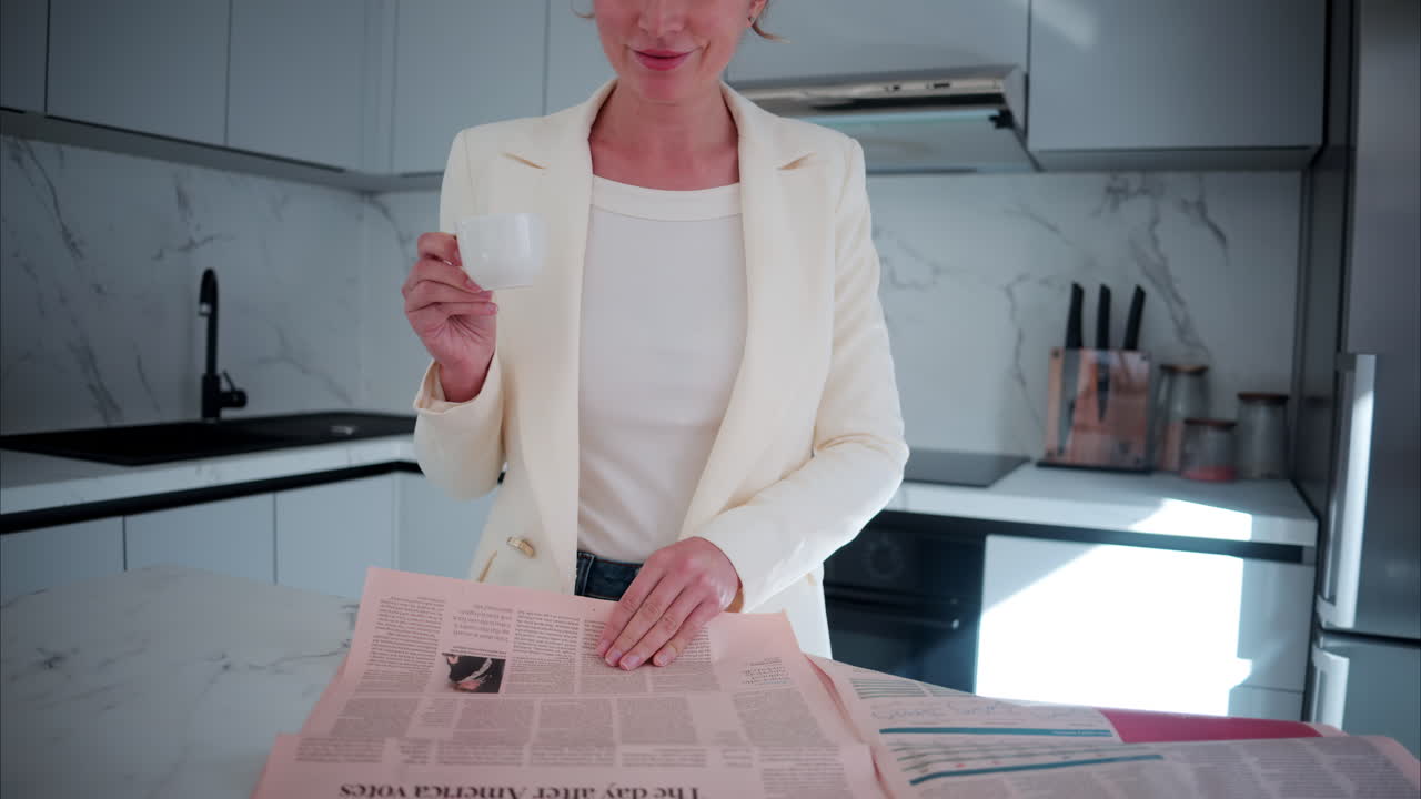 Donna in blazer bianco che sorseggia un caffè mentre legge il giornale in cucina