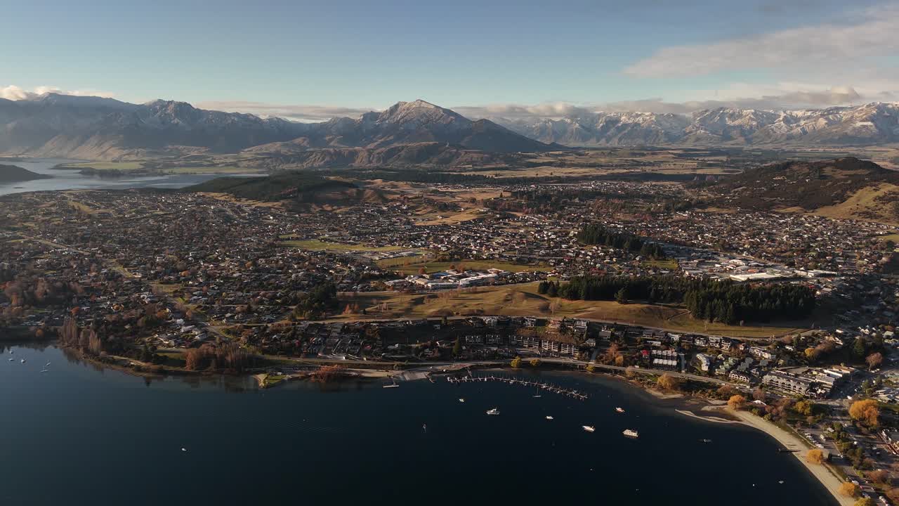 Wide panoramic aerial of Wanaka town on lakefront with snow-capped Southern Alps mountain range in background, New Zealand