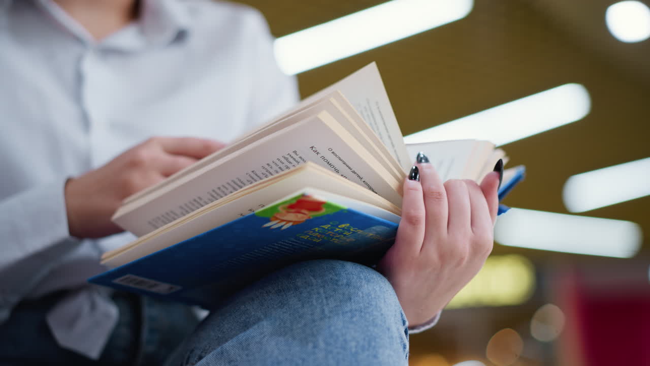 primer plano de una mujer con uñas negras volviendo páginas de un libro mientras está sentada con las piernas cruzadas, las suaves luces bokeh en el fondo crean una atmósfera cálida y moderna