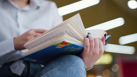 close-up of woman with black nails flipping through pages of a book while seated with crossed legs, soft bokeh lights in the background create a warm and modern atmosphere