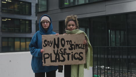 Zwei-Junge-Klimaaktivistinnen-Halten-Ein-Banner-Und-Protestieren-Gegen-Die-Einwegkunststoffe,-Während-Sie-In-Die-Kamera-Schauen