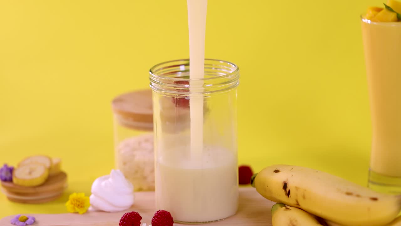 Versa un frullato di banana in un bicchiere con banane, lamponi e dolci su sfondo giallo acceso