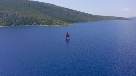 Luftaufnahme:-Verfolgung-Eines-Segelboots-Mit-Roten-Segeln,-Das-Im-Sommer-In-Der-Nähe-Der-Küste-Einer-Griechischen-Insel-Segelt