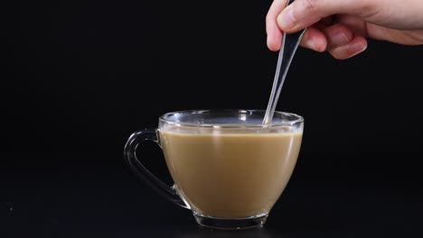 a hand stirs coffee with milk in a glass cup against a black background, highlighting the swirling motion