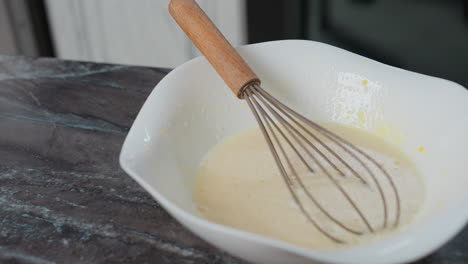 close-up of whisk resting in baking mixture inside white bowl on countertop, kitchen background features modern cabinets, sleek design, and clean workspace