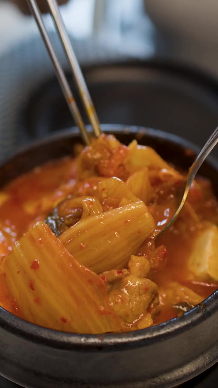 Bidikan jarak dekat sumpit logam mengaduk kimchi jjigae pedas dalam panci batu
